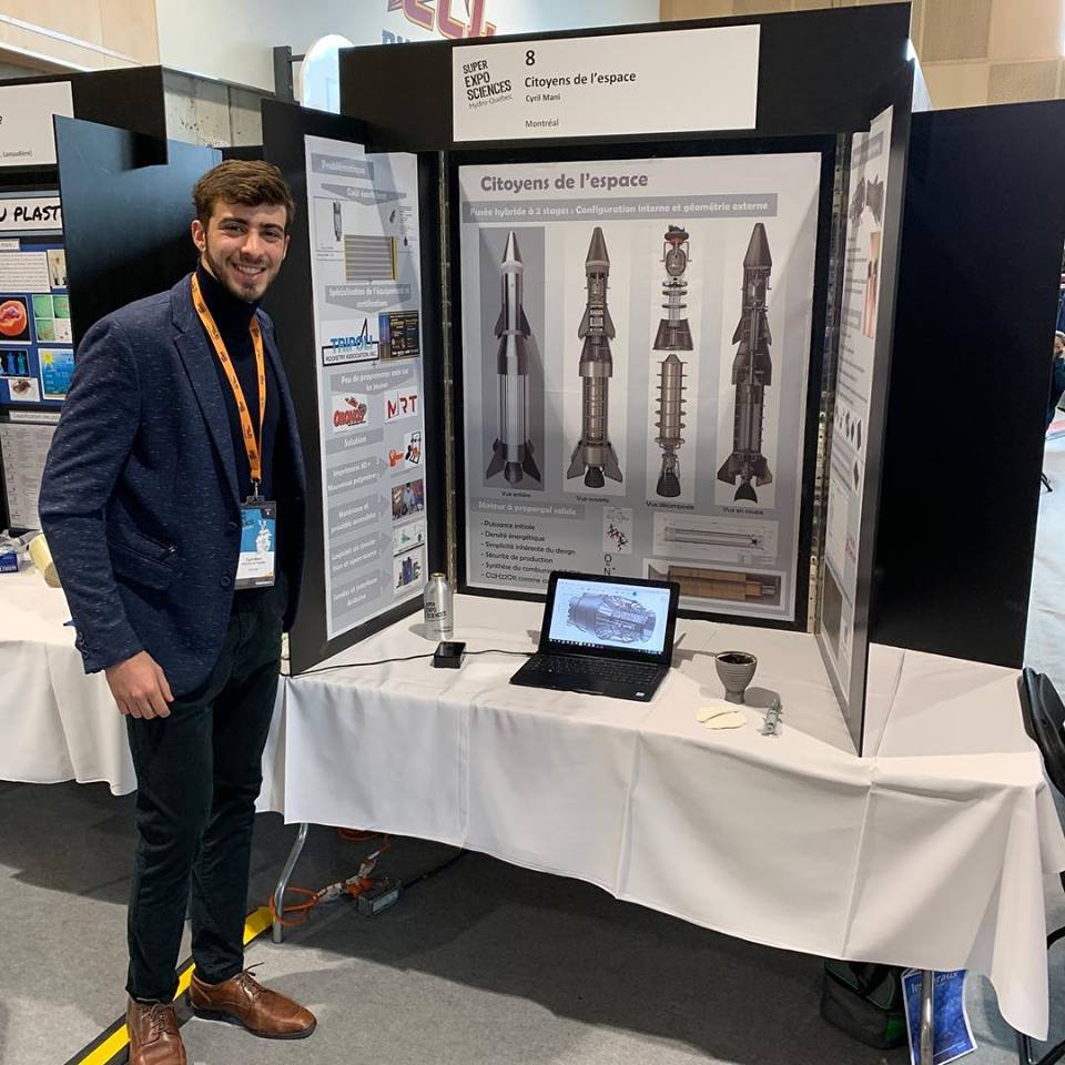 Cyril est devant son stand avec des affiches sur lesquelles sont dessinées de fusées. Sur la table un ordinateur portable.